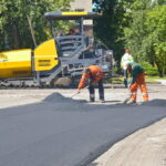 Ulica pokryta nową nawierzchnią bitumiczną. Widoczne sa znaki poziome. Po obu stronach ulicy domy i drzewa.