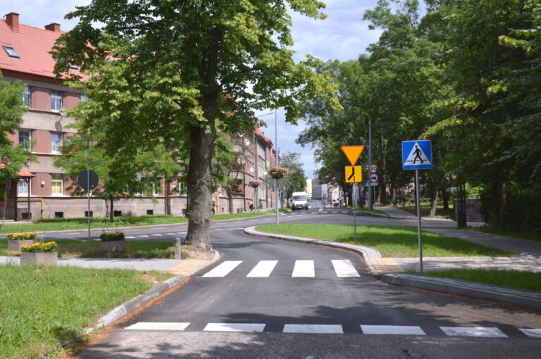 Ulica pokryta nową nawierzchnią bitumiczną. Widoczne sa znaki poziome. Po obu stronach ulicy domy i drzewa.