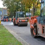 Maszyny drogowe układają nową nawierzchnię. Po obu stronach ulicy domy i drzewa.