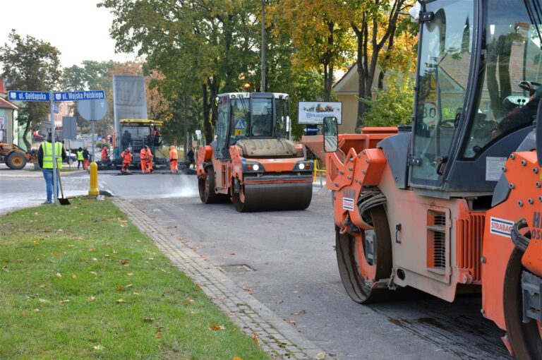 Maszyny drogowe układają nową nawierzchnię. Po obu stronach ulicy domy i drzewa.