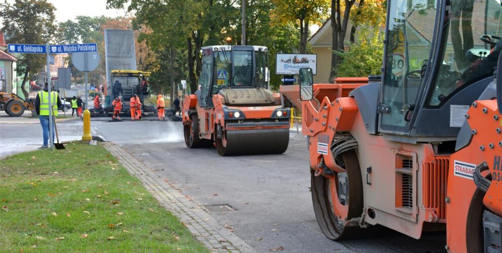 Maszyny drogowe układają nową nawierzchnię. Po obu stronach ulicy domy i drzewa.