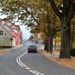 Nowa nawierzchnia bitumiczną. Widoczne sa znaki poziome. Po obu stronach ulicy domy i drzewa.