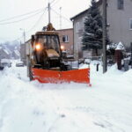 Ulica pokryta sniegiem. W traktor z pługiem do odśnieżania. Po obu stronach domy.
