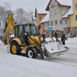 Ulica pokryta sniegiem. W traktor z pługiem do odśnieżania. Po obu stronach domy.