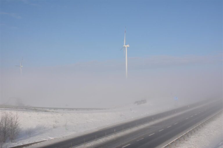 Odśniezona dwupasmowa droga. W głębi zamglona turbina wiatrowa.